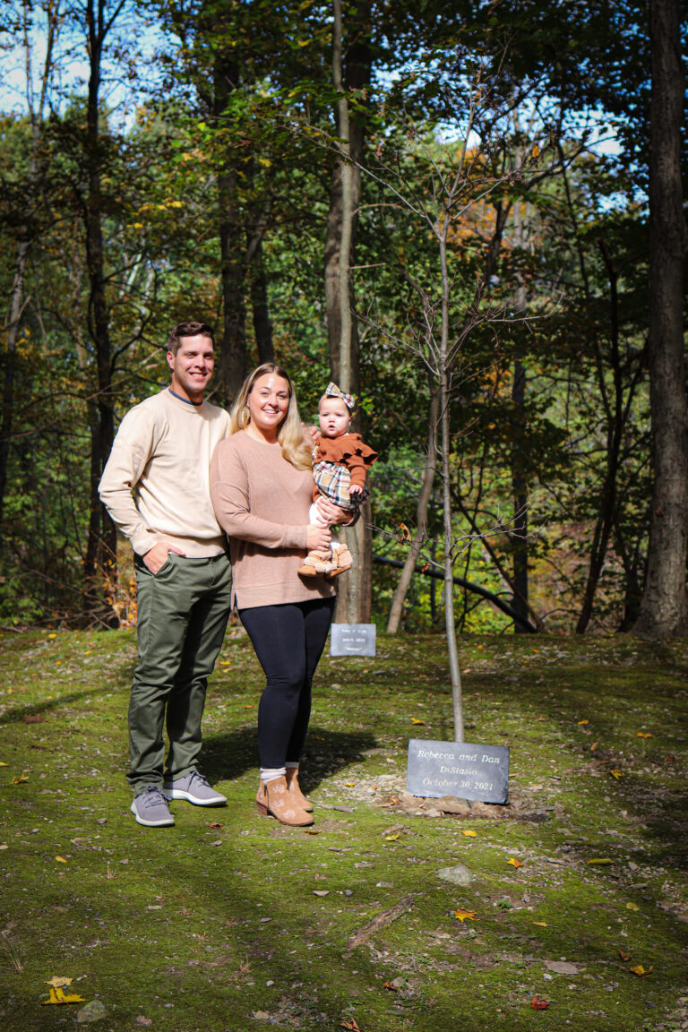 Mother and father stand near infant tree holding little girl